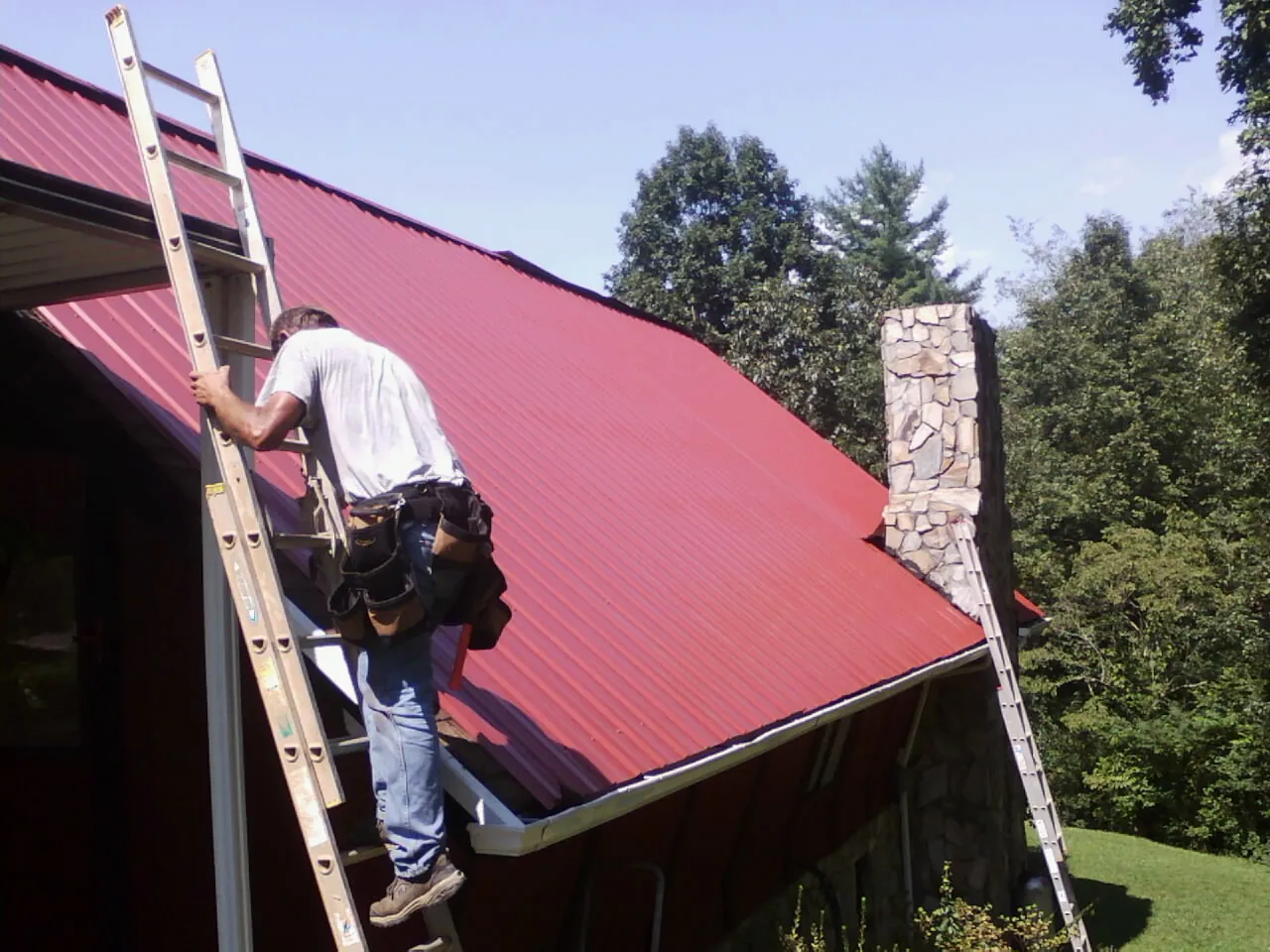 Roofer on a ladder beside a red metal roof and stone chimney — Soffit & Fascia Repair by Roofing Brothers in Myrtle Beach